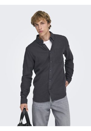 Camicia grigia scura con bottoni e taschino sul petto, maniche lunghe, realizzata in tessuto morbido, abbinata a jeans grigio chiaro, tenuta con una borsa nera.