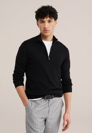 Muž s kudrnatými vlasy, který má na sobě černý svetr na zip přes bílou košili a šedé kalhoty s tenkými pruhy, stojí s jednou rukou v kapse.