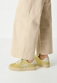 Sneakers gialli con superfici testurizzate, dettagli grigio chiaro e lacci color crema. Indossati con pantaloni a gamba larga beige chiaro. Design a suola piatta.