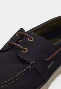 Ναυτικό μπλε παπούτσι τύπου boat με καφέ δερμάτινα κορδόνια, ορειχάλκινα κρίκους, λευκή ραφή και μπεζ σόλα, με μια μικρή ετικέτα με το λογότυπο Barbour.