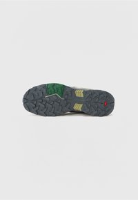Trail shoe outsole featuring a rugged gray rubber sole with green and yellow accents, textured tread for traction, and a red triangle detail.