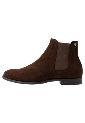 Botte Chelsea en daim marron avec panneau latéral élastique, talon bas, bout rond et détail de logo métallique sur la languette du talon arrière.