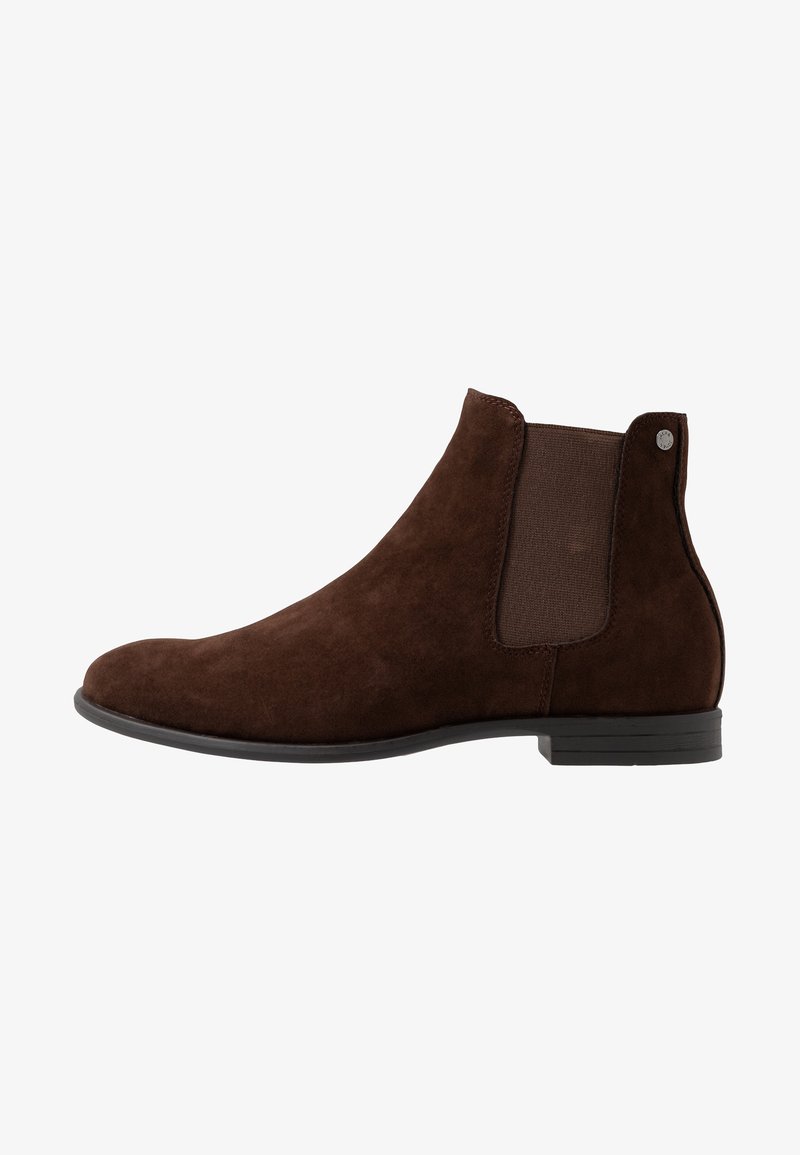 Botte Chelsea en daim marron avec panneau latéral élastique, talon bas, bout rond et détail de logo métallique sur la languette du talon arrière.