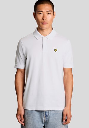 Mann trägt ein einfarbiges weißes Poloshirt mit einem kleinen gelben Vogel-Logo auf der linken Brust, kombiniert mit hellblauen Jeans.