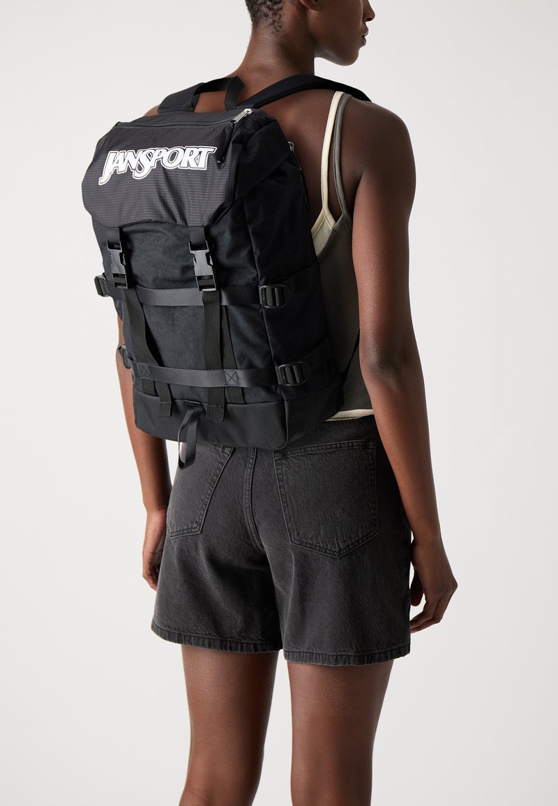 Sac à dos Jansport noir avec fermeture à glissière, bretelles réglables et plusieurs boucles à l'avant ; fabriqué en tissu durable avec une finition texturée.