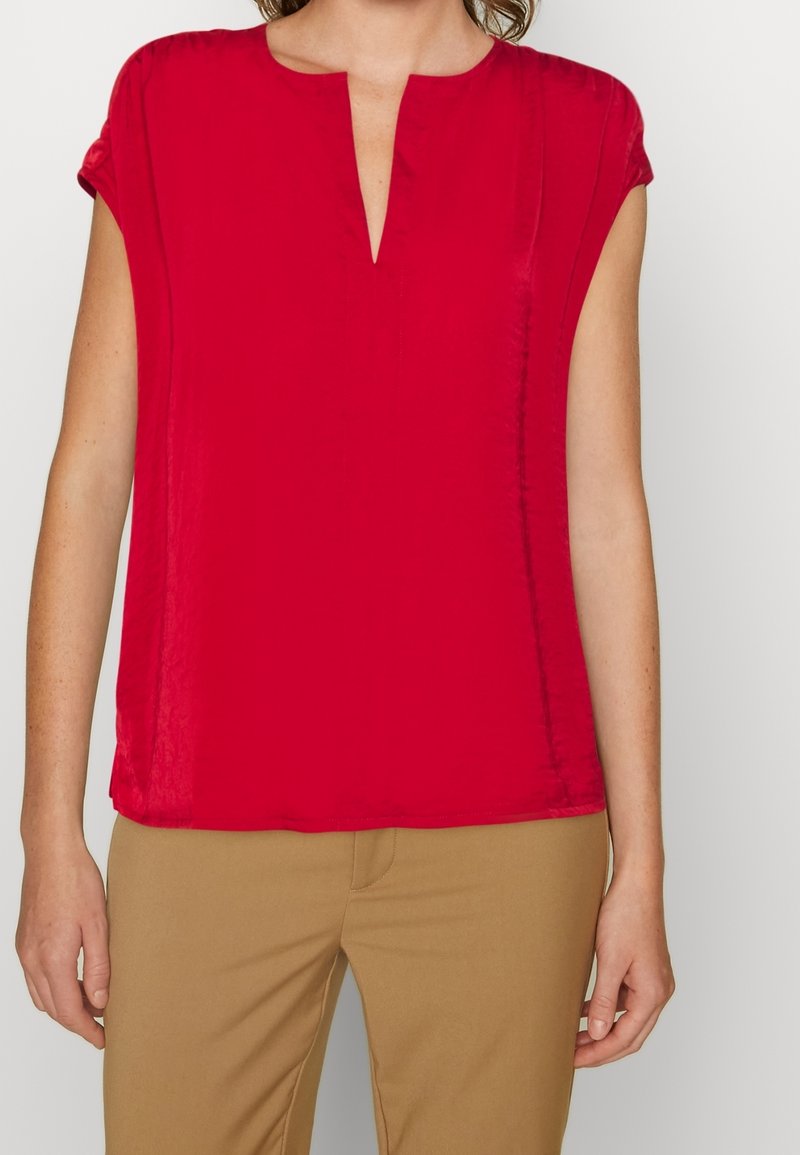 Blouse rouge à manches courtes avec un col en V, tissu lisse, coupe décontractée et fentes latérales, associée à un pantalon beige.