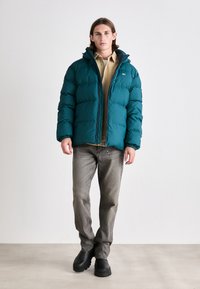 Teal puffer jacket with a front zipper and hood, featuring elastic cuffs. Worn over a beige shirt and paired with gray jeans and black boots.