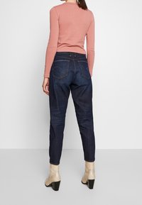 Jeans de mezclilla oscura con forma ajustada, presentan dos bolsillos traseros y costuras en contraste, combinados con una blusa de manga larga en tono rosa apagado y botas de tacón en color beige.