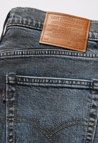 Blå denimjeans med en brun läderlapp med präglad text. Sömmen syns på fickor och sömmar och har klassiska designdetaljer.