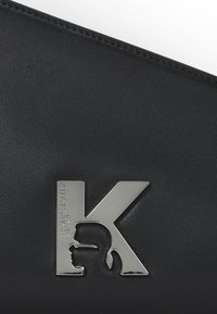 Svart läderplånbok med en silverfärgad metalllogotyp med bokstaven "K" och en silhuett. Slät yta med rena sömmar och minimal detaljering.