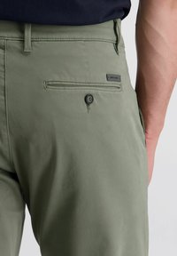 Olivgrüne Hosen aus weichem Stoff, mit einer geknöpften Gesäßtasche und einem Markenetikett. Das Design hat eine glatte Textur.