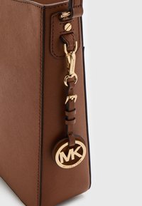 Bruine gestructureerde handtas met een getextureerde afwerking, voorzien van gouden beslag en een ronde logohanger bevestigd aan een leren riem.