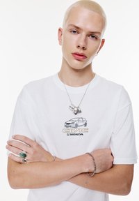 Witte t-shirt met een autografiek en de tekst "CIVIC", zilveren ketting met hanger, groene stenen ring en zilveren armband op gekruiste armen.