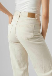 Beige Jeans aus Denim mit hoher Taille, zwei Gesäßtaschen und einem braunen Lederpatch am Bund. Texturiert.