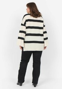 Gestreifter Pullover in Schwarz-Weiß mit weiten Ärmeln, weichem Material und unregelmäßigem Saum, kombiniert mit schwarzen Hosen und Stiefeln mit Absatz.