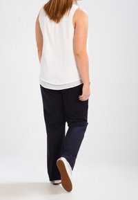 Haut blanc sans manches à texture lisse, associé à un pantalon bleu marine. Baskets blanches à semelles marron vues de dos.