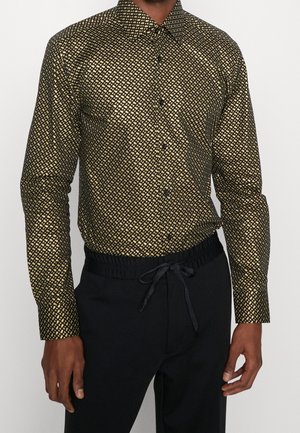 Homme portant une chemise à manches longues avec un motif géométrique doré et noir, rentrée dans un pantalon noir à cordon.