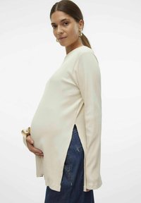 Kreminė pregmatinė viršūnė su ilgomis rankovėmis, šoninėmis plyšėmis ir laisvu pritaikomu stiliumi, vilkima ant tamsiai mėlynų džinsų. Minkšta tekstūra ir kasdienis dizainas.