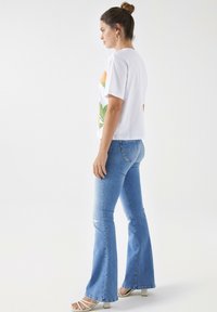 Camiseta blanca de manga corta con estampado floral, combinada con jeans acampanados de color azul claro. El modelo lleva sandalias de tacón blancas.