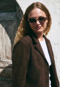 Blazer en daim marron avec un col structuré et une fermeture à un bouton sur un T-shirt blanc. Le mannequin porte des lunettes de soleil à pois et a des cheveux ondulés.