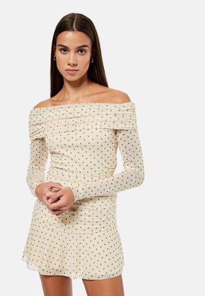 Pitkätukkainen ruskeahiuksinen nainen, jolla on päällä olkapäät paljastava beige mekko mustilla pilkuilla, seisoo kätensä puuskassa yksiväristä taustaa vasten.