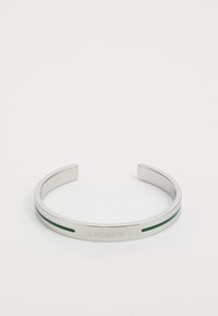 Ezüst színű fém karkötő, zöld díszcsíkkal, amelyen a 'LACOSTE' felirat található. Sima textúrával, mindkét végén nyitott dizájnnal.