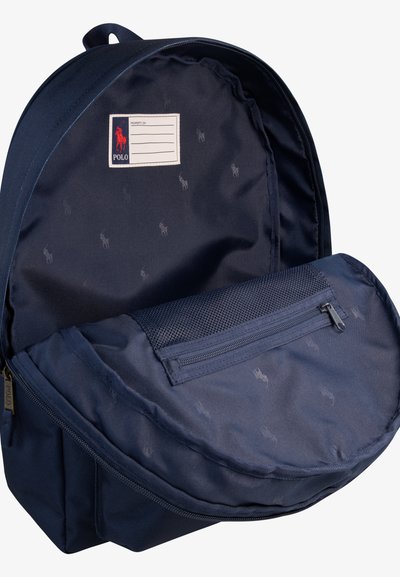 Marinblå ryggsäck med mjukt tyg, utrustad med en framficka med dragkedja och en etikettplats. Insidan visar ett subtilt mönster av logotyper. Rund form.