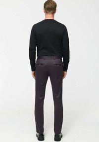 Man wearing black long-sleeve shirt, dark purple tailored pants with back pockets, and black shoes standing facing away on white background.