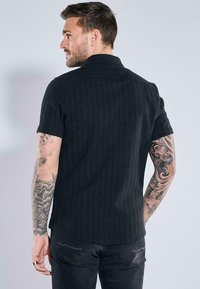 Schwarzes Kurzarmhemd mit einem strukturierten, gerippten Design; verfügt über einen klassischen Kragen und einen abgerundeten Saum. Das Modell zeigt das Hemd von hinten.
