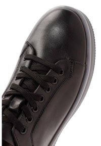 Zapatilla negra de cuero con punta redondeada, agujetas negras y superficie texturizada. Presenta una suela de goma blanca contrastante.