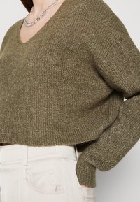 Pull court en maille vert olive avec un décolleté en V, tissu texturé et manches oversize, associé à un pantalon taille haute de couleur claire.