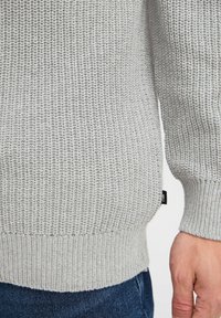 Suéter de punto gris claro con un patrón texturizado, puños y dobladillo acanalados, con una etiqueta lateral. Adecuado para vestir de manera casual.