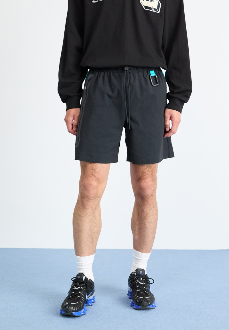 Homme portant un short de sport noir avec un mousqueton accroché à la ceinture, des chaussettes blanches et des chaussures de course noires à semelles bleues.