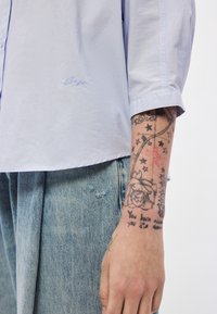 Ανοιχτό μπλε πουκάμισο με κοντά μ�ανίκια και διακριτικό κεντημένο λογότυπο, φορεμένο με ανοιχτόχρωμο τζιν παντελόνι που διαθέτει φθαρμένη υφή και εμφανείς ραφές.