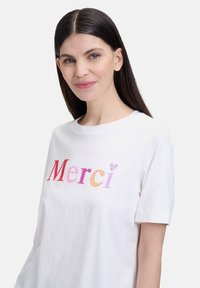 White cotton t-shirt with short sleeves featuring a multicolored "Merci" text design, using red, orange, pink, and lavender hues.