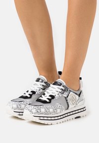 Sneaker in bianco e nero con tomaia in rete e motivo, dotate di suola testurizzata, lacci bianchi, borchie decorative e un dettaglio del logo.