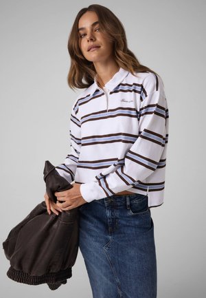 Modèle portant un polo blanc à manches longues et à rayures ainsi qu’une jupe en jean bleue, tenant une veste en cuir marron foncé devant un fond de studio gris.