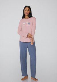 Top de manga larga color rosa claro con gráfico de corazón y texto "Mantén la Felicidad", combinado con pantalones azules holgados con dobladillos con volantes. Material de algodón suave.