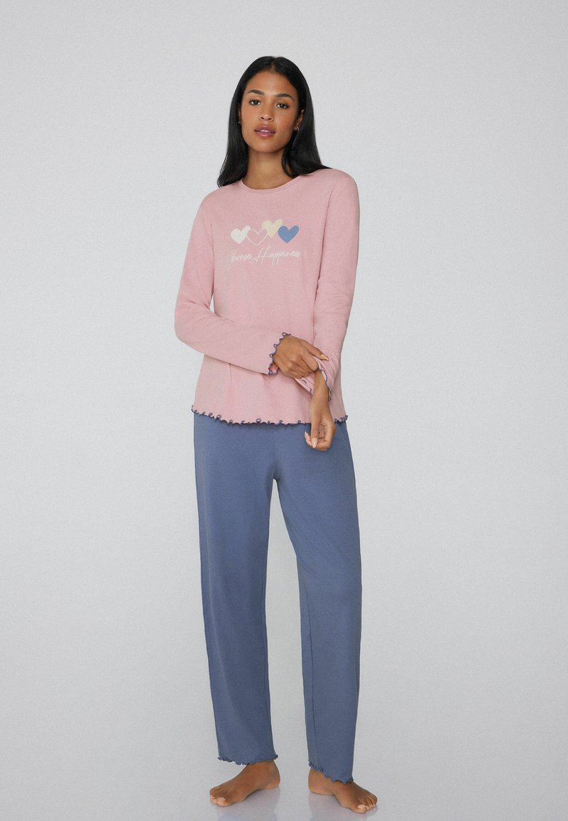 Top de manga larga color rosa claro con gráfico de corazón y texto "Mantén la Felicidad", combinado con pantalones azules holgados con dobladillos con volantes. Material de algodón suave.