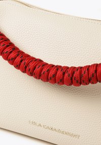 Off-white skórzana torba o teksturowanej powierzchni, z czerwoną plecioną rączką z czarnymi akcentami oraz złotym logo na przedzie.