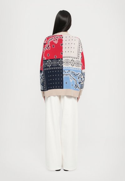 Cardigan en tricot épais rouge, bleu marine, beige et bleu clair avec des motifs bandana. Présente une coupe décontractée et un ourlet côtelé, associé à un pantalon large blanc.