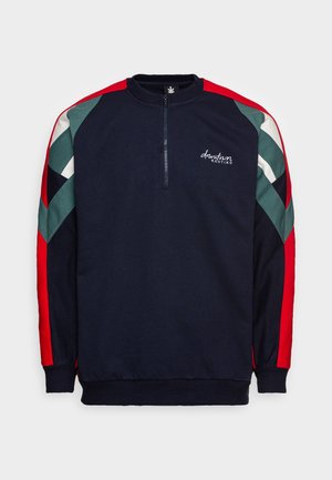 Jersey azul marino con franjas en forma de cheurón rojas, verdes y blancas en las mangas, cremallera corta frontal, puños y bajo acanalados, y texto "downtown KAOTIKO" en el pecho.