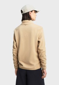 Veste en polaire beige avec un col montant, texture douce et poignets élastiques, accompagnée d'une casquette de couleur claire visible en arrière-plan.