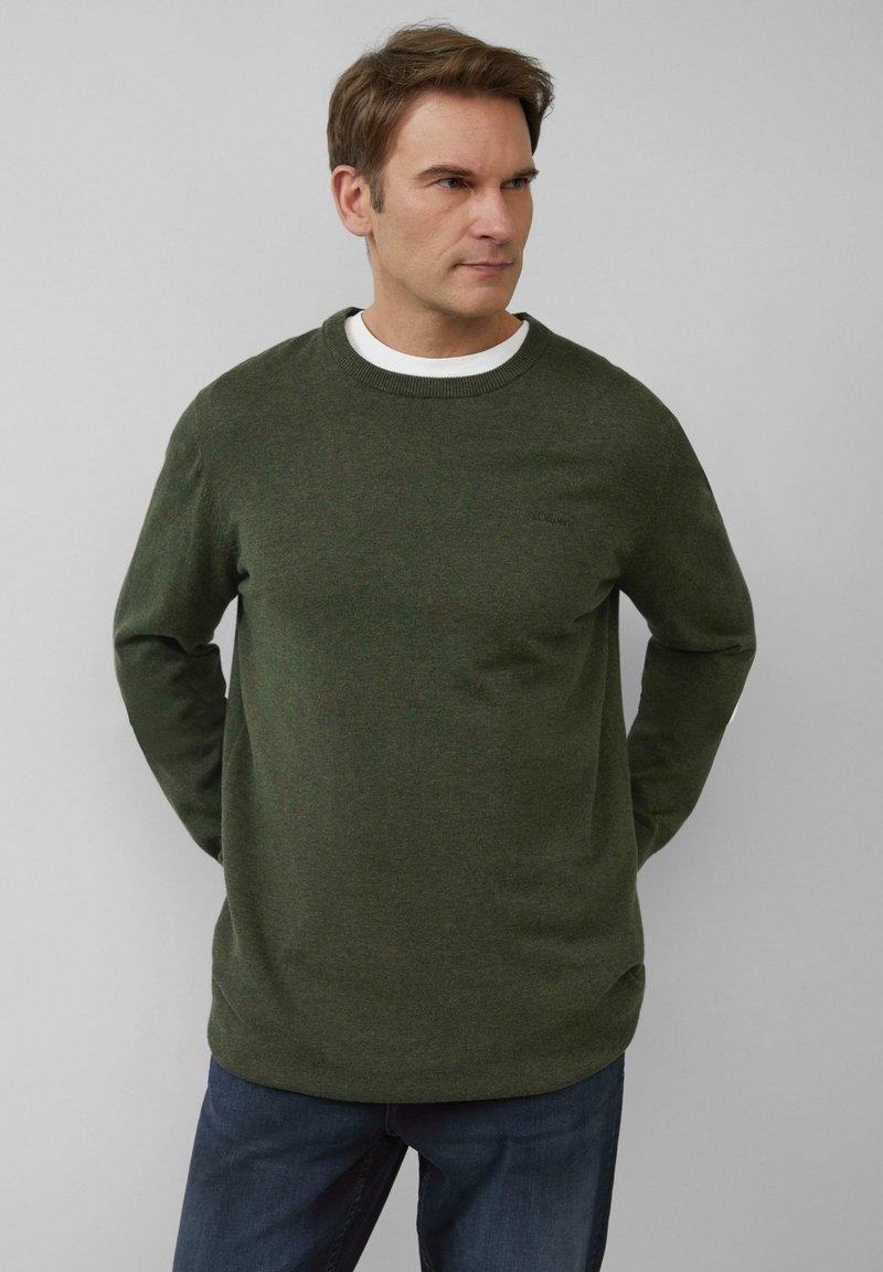 Dunkelgrüner Strickpullover mit langen Ärmeln, Rundhalsausschnitt und abgerundetem Saum. Verfügt über ein kleines Markenlogo auf der Brust. Wird über einem weißen Hemd getragen.