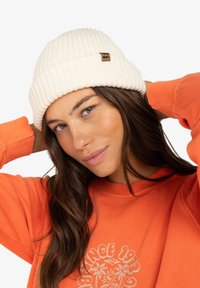 Witte geribbelde gebreide beanie met een bruine leren logo patch. Draagt samen met een oranje sweatshirt met een grafisch ontwerp. Zachte textuur.