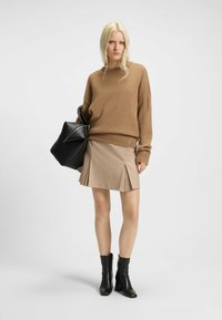 Brauner Strickpullover, beiger Faltenrock mit Nadelstreifen und schwarze Absatzstiefeletten mit Knöchelschnitt. Modell hält eine schwarze Tasche mit kantigem Design.