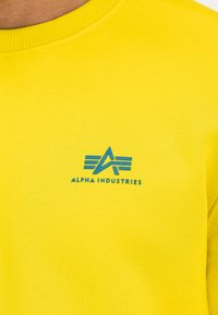 Gul genser med ribbet hals, med en blå logo "ALPHA INDUSTRIES" og et geometrisk mønster på forsiden.