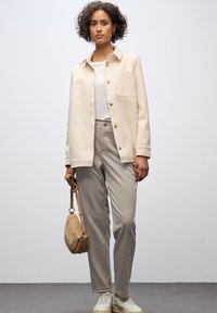 Beige overskjorta i mjukt material, kombinerad med ljusgrå skräddarsydda byxor. Modellen håller en brun handväska och har på sig vita sneakers.