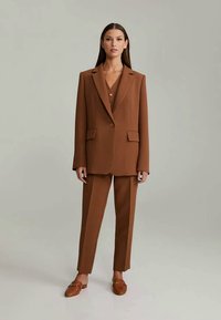 Hnědý tailored blazer a odpovídající kalhoty ve strukturovaném designu, s jedním knoflíkem a bočními kapsami, v kombinaci s hnědými slip-on botami.