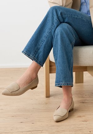 Person iført beige lave hælede loafers og blå jeans, siddende på en lys træstol med en beige pude på et trægulv.